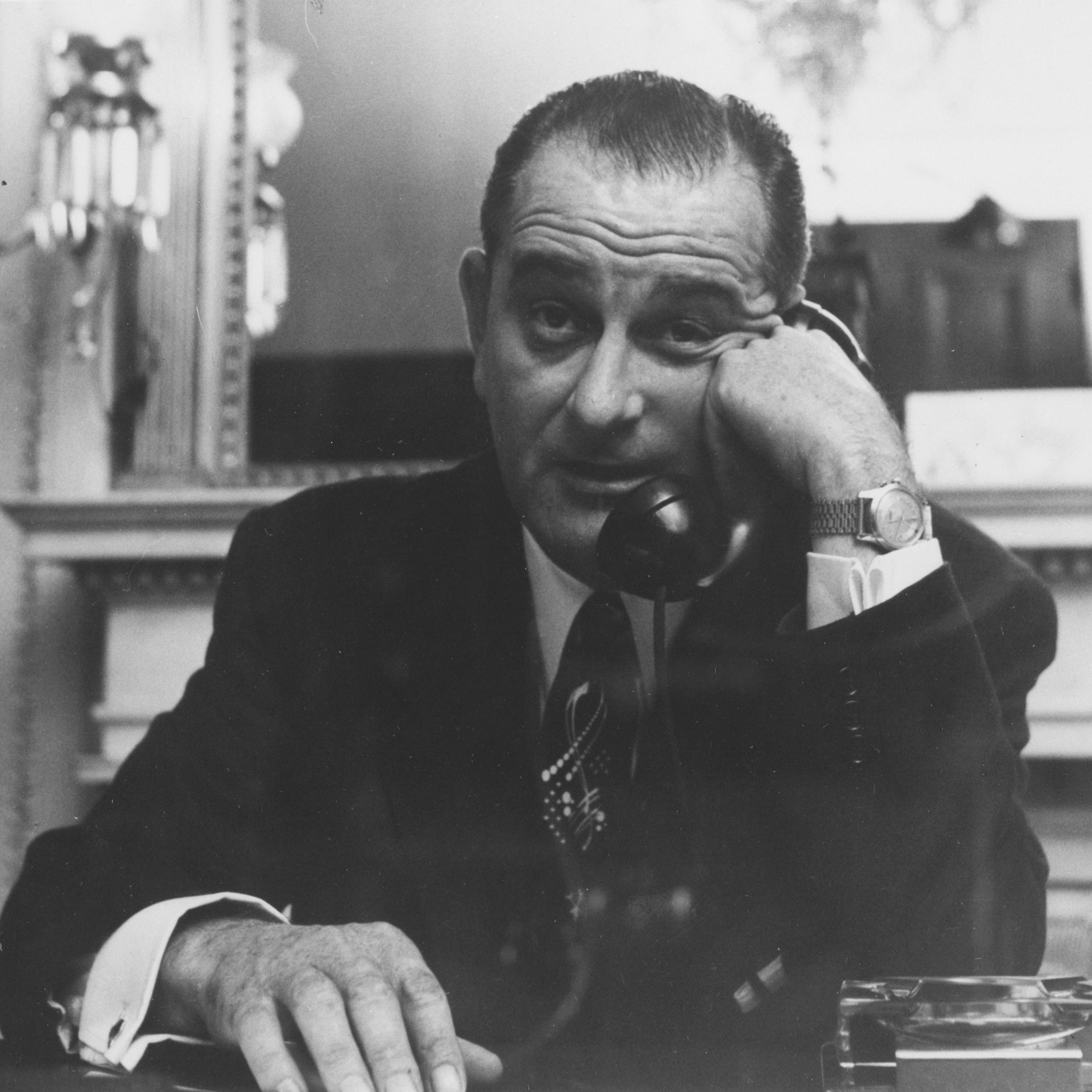 Lyndon Baines Johnson raises his eyebrows as he talks on the phone, seated behind a desk. 